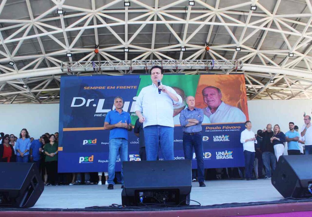 Gilberto Kassab discursando em convenção política com Dr. Luiz Braz ao fundo, promovendo a reeleição em Campo Limpo Paulista.