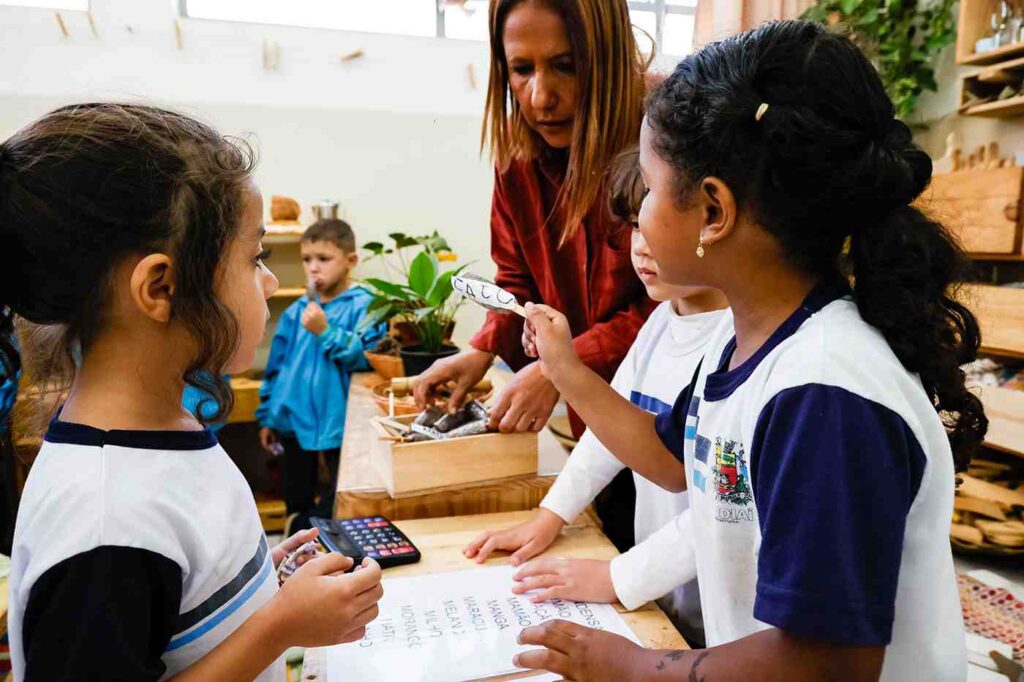 Professora ajudando crianças com atividades de alfabetização em Jundiaí na EMEB Cleo Nogueira Barbosa, interação e aprendizado.