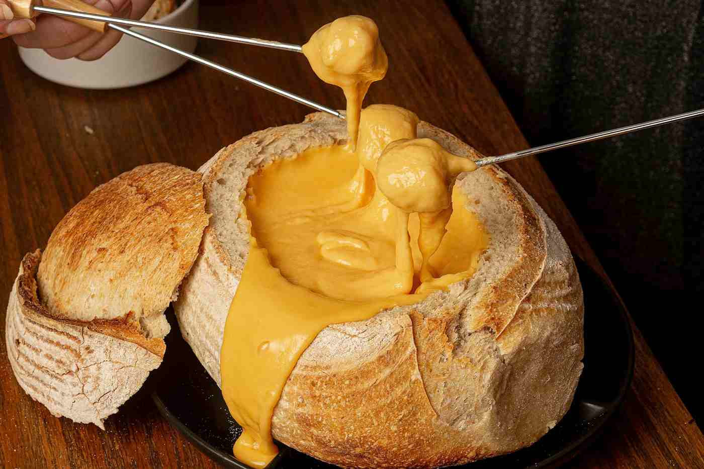 fondue de queijo servido dentro de um pão rústico, com garfos de fondue mergulhando pedaços no queijo derretido.