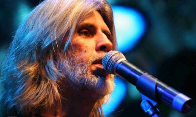 Osvaldo Montenegro cantando no microfone em um show, com cabelo e barba grisalhos, iluminação azul de fundo.