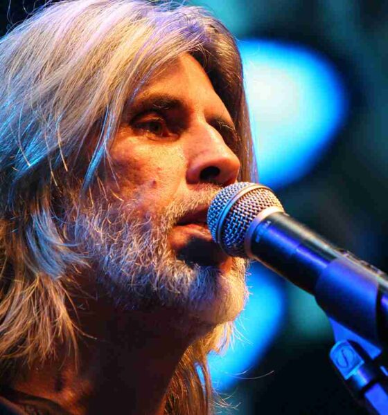 Osvaldo Montenegro cantando no microfone em um show, com cabelo e barba grisalhos, iluminação azul de fundo.
