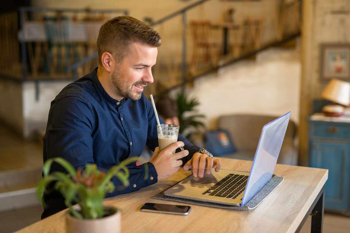 Homem sorridente trabalhando em um laptop em um café, bebendo uma bebida gelada; ambiente casual ideal para pequenas empresas.