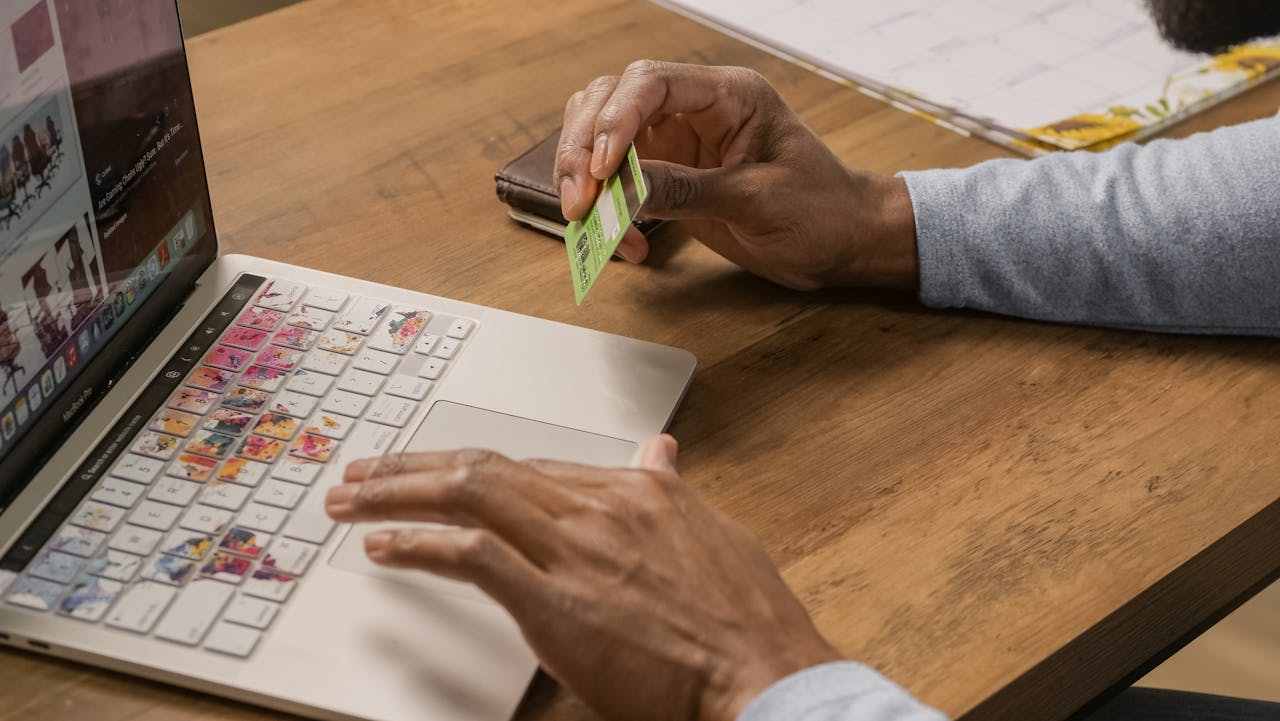 Homem segurando cartão de crédito verde e usando laptop em mesa de madeira, ilustrando compras online e uso consciente de crédito.