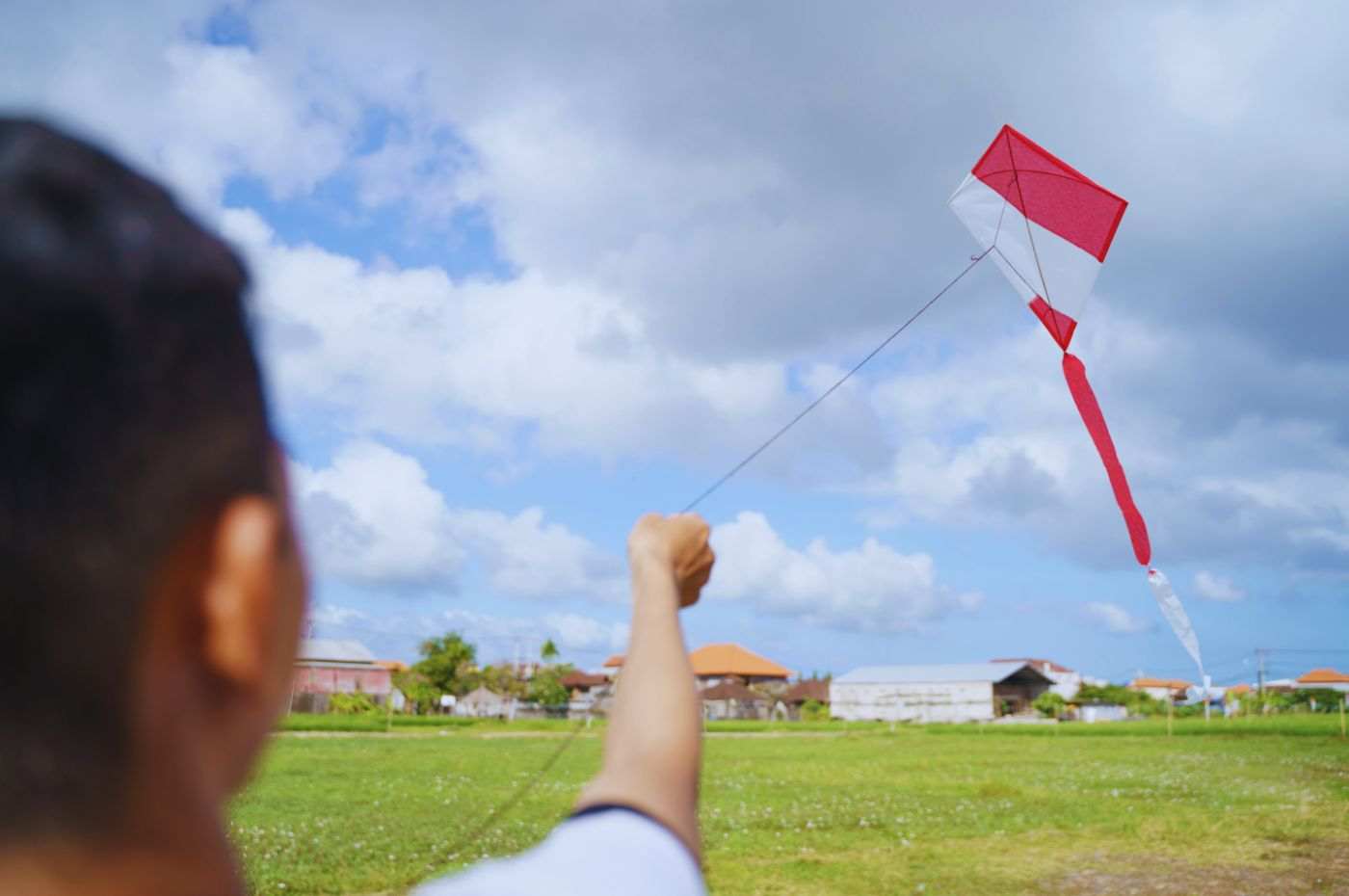 Pessoa empinando pipa vermelha e branca em campo aberto, evitando interrupções de energia perto de redes elétricas.