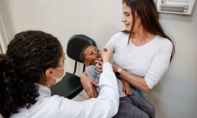Mãe segurando criança enquanto profissional de saúde aplica Vacina contra a Paralisia Infantil em UBS