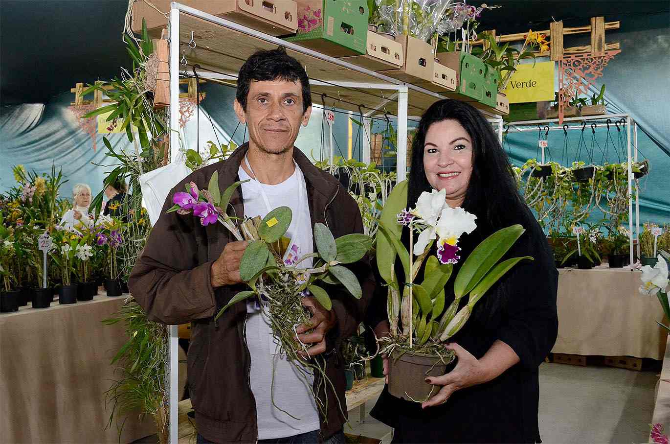 Casal exibe orquídeas na Festa das Orquídeas 2024, rodeados por diversas plantas em exposição no Clube de Campo da Cica.