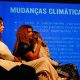 Mulheres discutem políticas para crianças e mudanças climáticas em painel, com apresentação em tela ao fundo destacando os riscos.