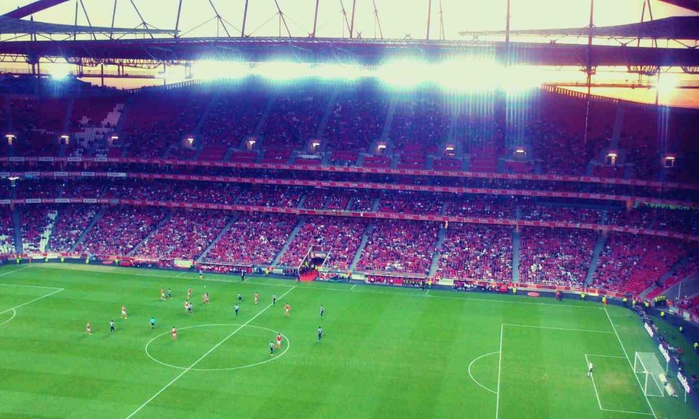 Estádio de futebol iluminado ao entardecer, com jogadores em campo durante uma partida, enquanto o VAR é utilizado para revisões.