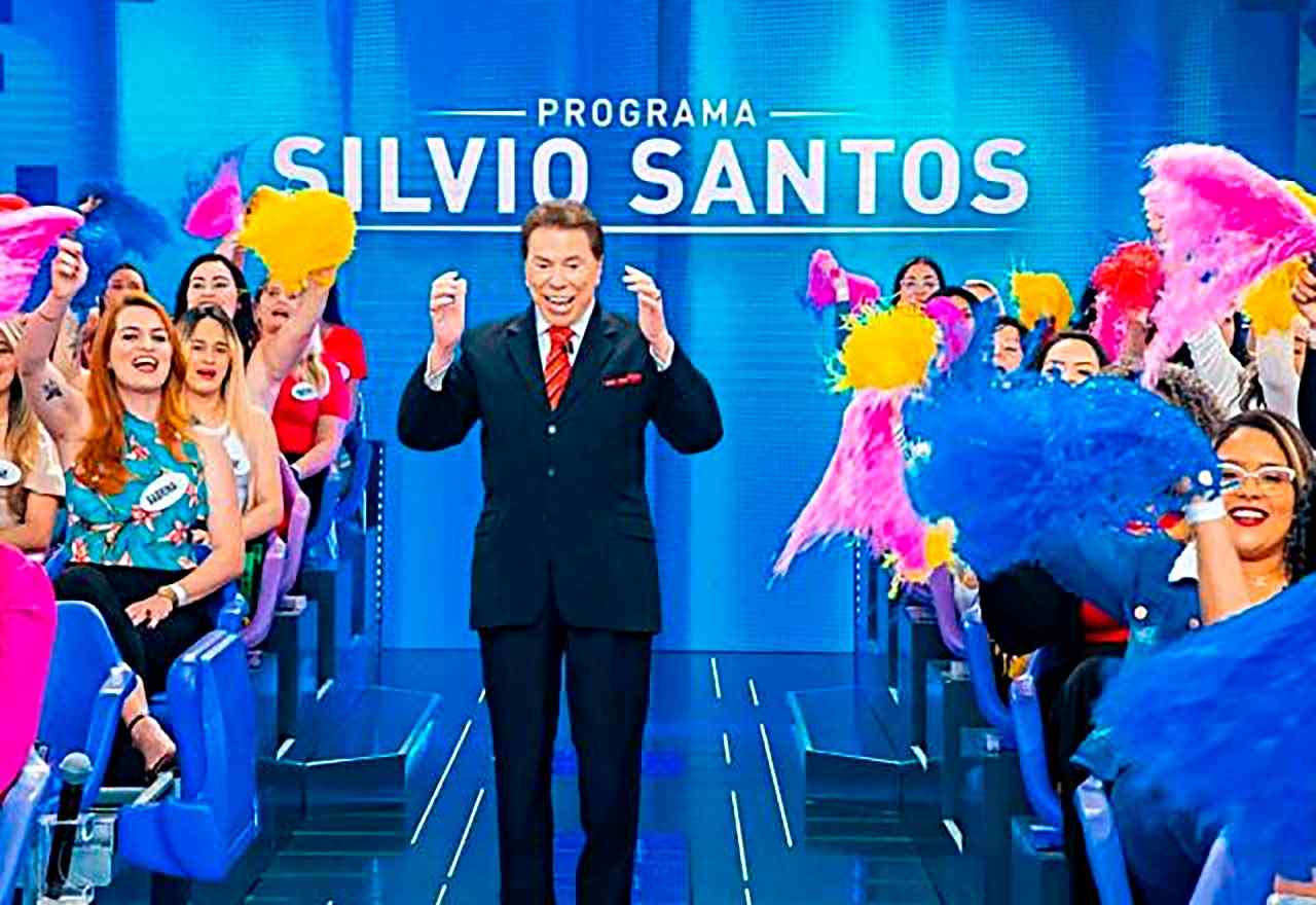 Silvio Santos sorridente no palco do Programa Silvio Santos, cercado por uma plateia animada com pompons coloridos.