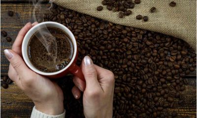 Mãos segurando uma xícara de café quente sobre uma superfície com grãos de café espalhados e saco de juta ao fundo.