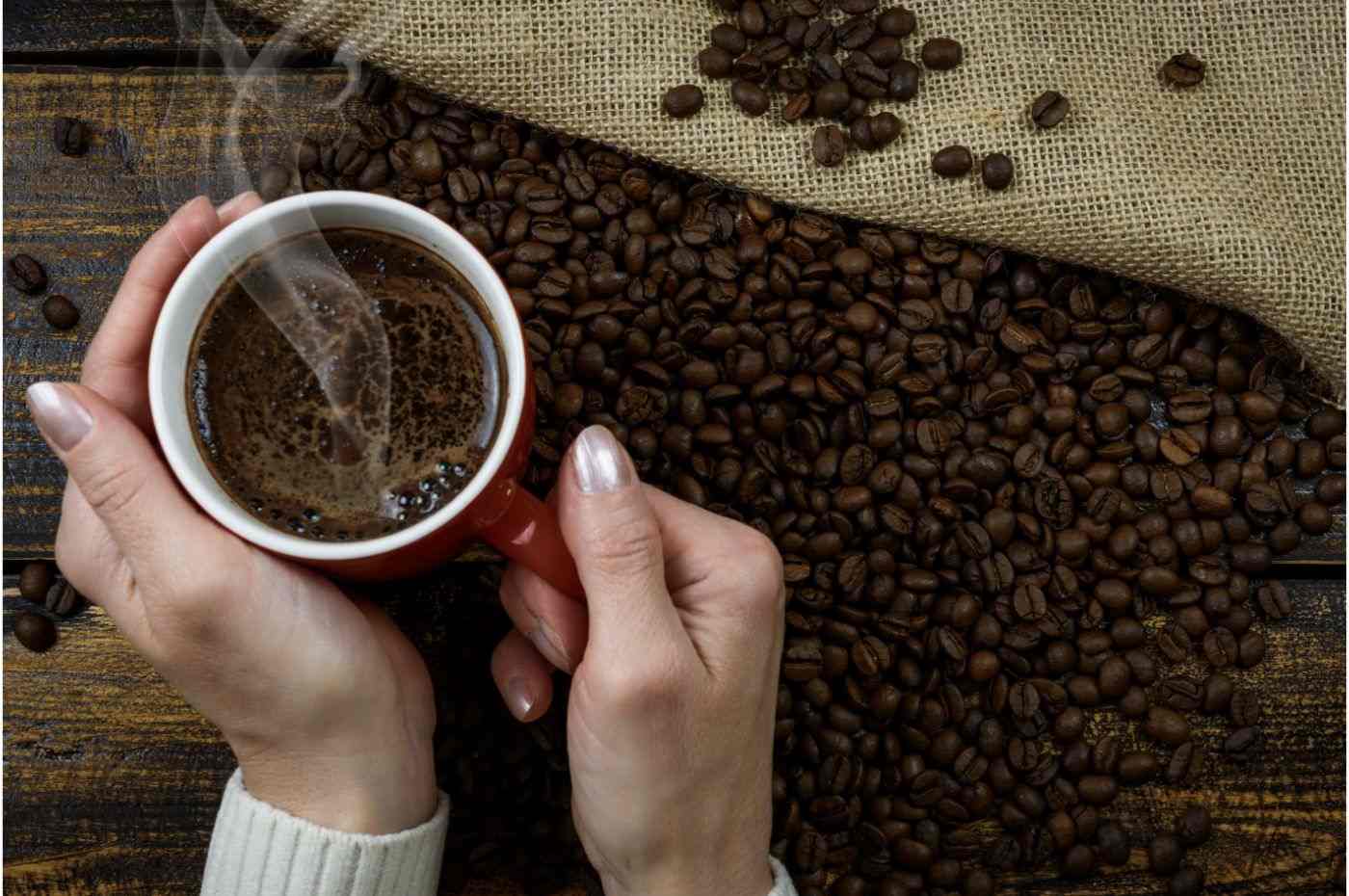 Mãos segurando uma xícara de café quente sobre uma superfície com grãos de café espalhados e saco de juta ao fundo.