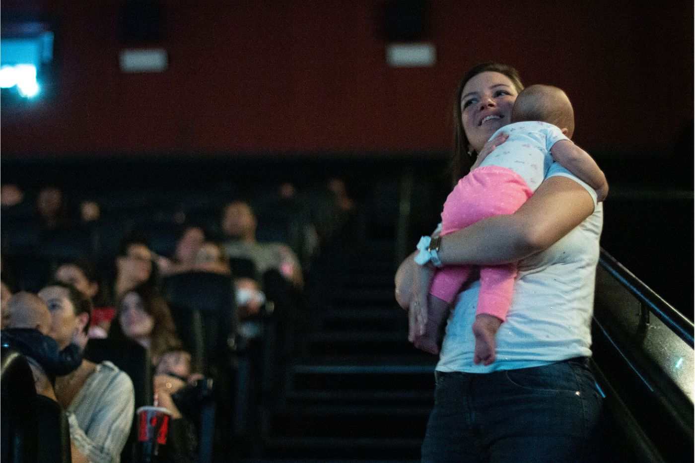 Mulher com bebê no colo assiste a sessão CineMaterna no cinema, ambiente acolhedor e adaptado para famílias com crianças