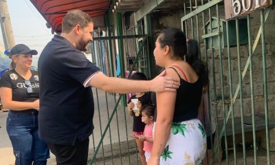 João Paulo cumprimenta moradora em frente a uma casa em Várzea Paulista durante caminhada, acompanhado por apoiadora e crianças ao fundo.