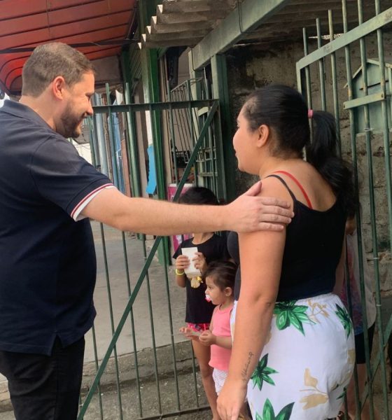 João Paulo cumprimenta moradora em frente a uma casa em Várzea Paulista durante caminhada, acompanhado por apoiadora e crianças ao fundo.