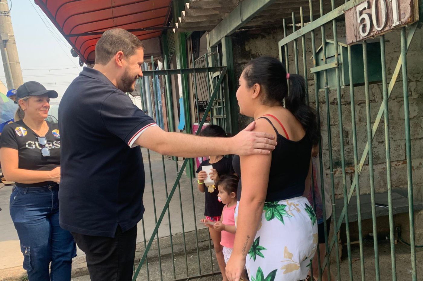 João Paulo cumprimenta moradora em frente a uma casa em Várzea Paulista durante caminhada, acompanhado por apoiadora e crianças ao fundo.