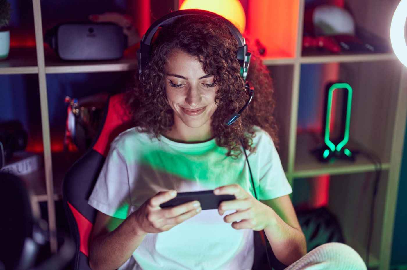 Mulher sorrindo jogando em cassino online no celular, com fones de ouvido e ambiente iluminado, refletindo a atmosfera de jogos.