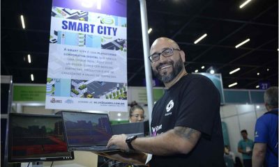 Homem sorridente do Grupo iv2 segura laptop em estande da Feira do Empreendedor, destacando a plataforma de realidade virtual Smart City.