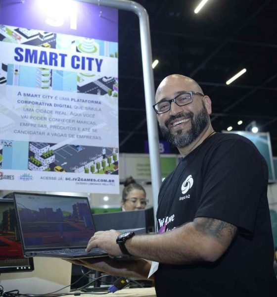 Homem sorridente do Grupo iv2 segura laptop em estande da Feira do Empreendedor, destacando a plataforma de realidade virtual Smart City.