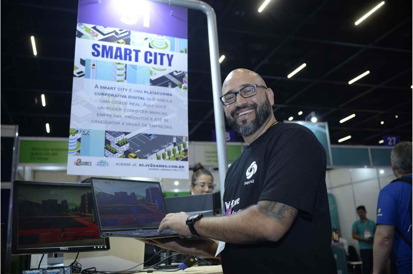 Homem sorridente do Grupo iv2 segura laptop em estande da Feira do Empreendedor, destacando a plataforma de realidade virtual Smart City.