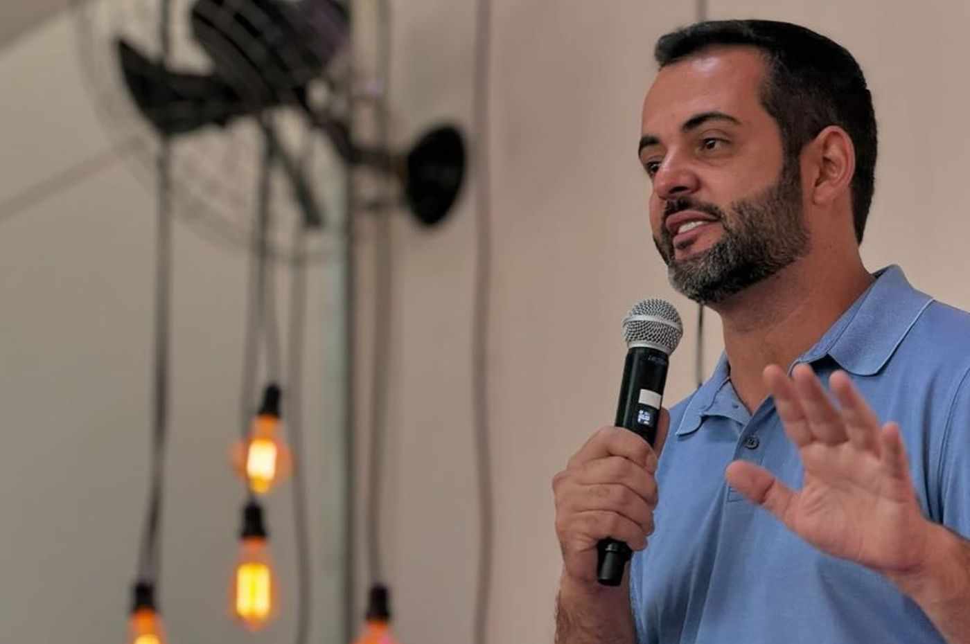 Gustavo Martinelli, homem com barba, usando camisa polo azul, segurando microfone e fazendo discurso, com lâmpadas penduradas ao fundo.