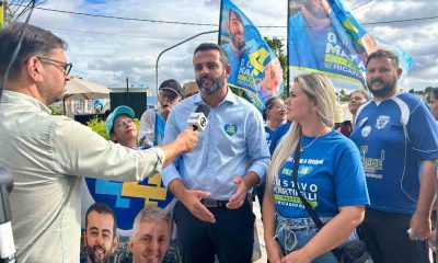 Gustavo Martinelli sendo entrevistado em evento de campanha na Vila Rio Branco, cercado por apoiadores com bandeiras.