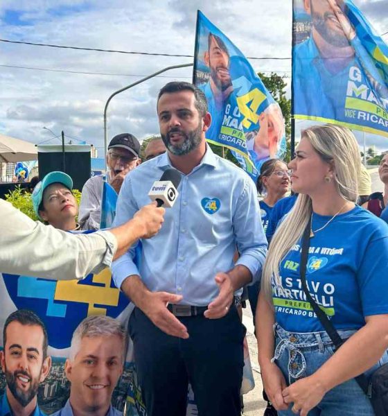 Gustavo Martinelli sendo entrevistado em evento de campanha na Vila Rio Branco, cercado por apoiadores com bandeiras.