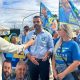 Gustavo Martinelli sendo entrevistado em evento de campanha na Vila Rio Branco, cercado por apoiadores com bandeiras.