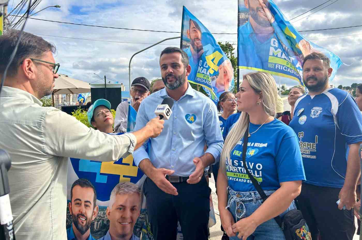 Gustavo Martinelli sendo entrevistado em evento de campanha na Vila Rio Branco, cercado por apoiadores com bandeiras.