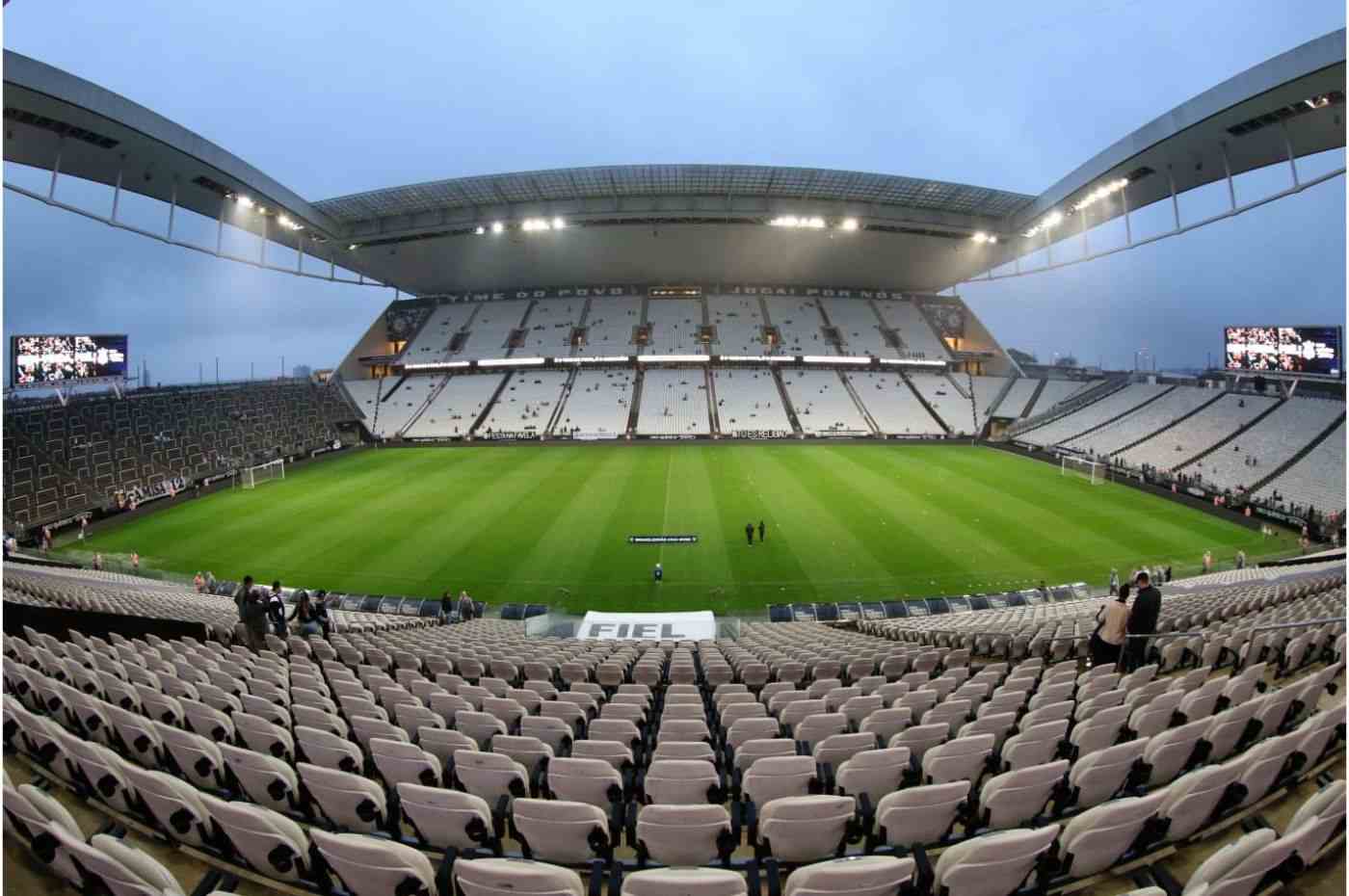 Estádio Neo Química Arena vazio com cadeiras brancas, gramado bem cuidado e iluminação ligada antes de uma partida de futebol.