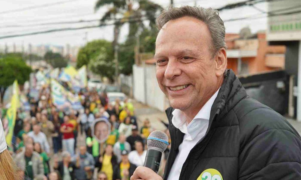 José Antonio Parimoschi segurando microfone e sorrindo durante caminhada de campanha em Jundiaí, cercado por apoiadores com bandeiras.