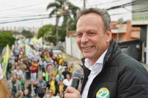 José Antonio Parimoschi segurando microfone e sorrindo durante caminhada de campanha em Jundiaí, cercado por apoiadores com bandeiras.