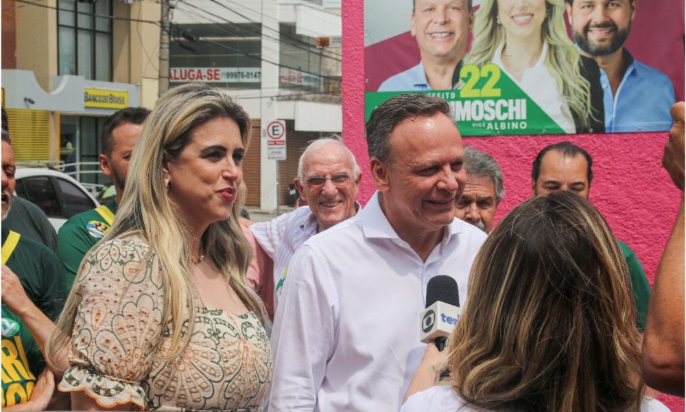 José Antonio Parimoschi e Quézia de Lucca durante campanha em Jundiaí, conversando com a imprensa em frente a um cartaz eleitoral