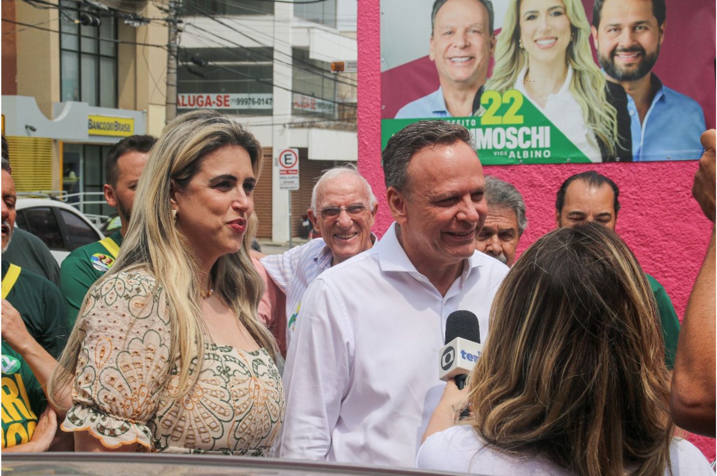 José Antonio Parimoschi e Quézia de Lucca durante campanha em Jundiaí, conversando com a imprensa em frente a um cartaz eleitoral