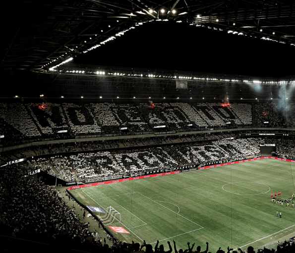 Torcida organizada exibe mosaico contra crimes em estádios, com mensagem de união e paz antes de partida de futebol em estádio iluminado.