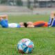 Dois jogadores descansam deitados no gramado após treino, com bola de futebol em destaque no campo. Data Fifa.