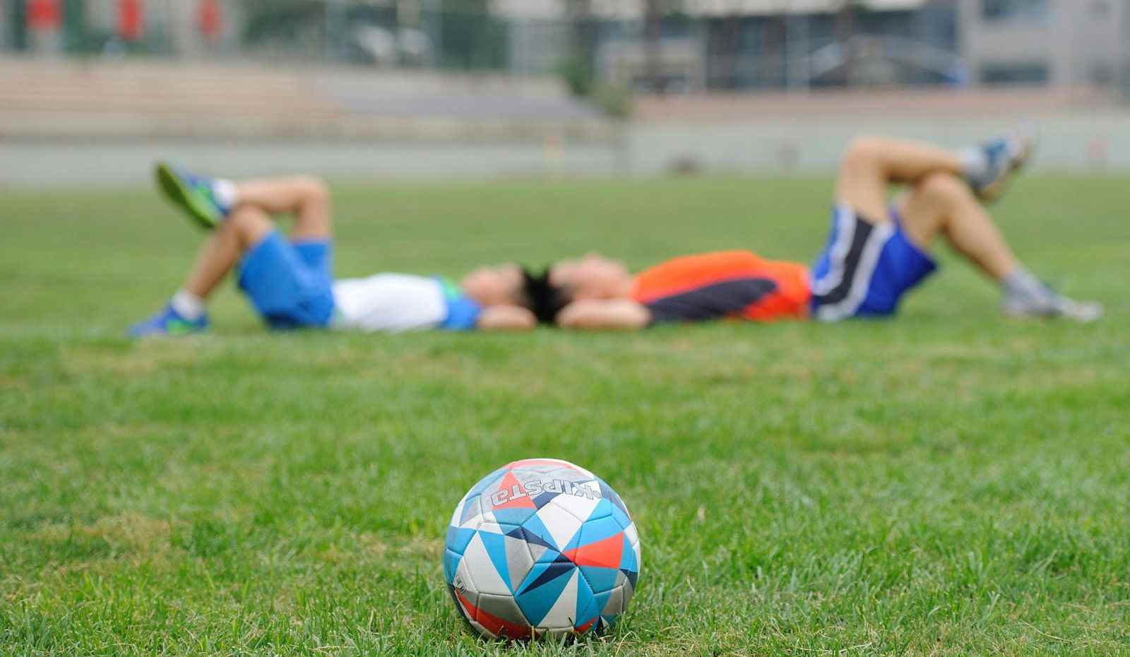 Dois jogadores descansam deitados no gramado após treino, com bola de futebol em destaque no campo. Data Fifa.