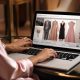 Mulher usando laptop para navegar em site de moda, visualizando opções de roupas e acessórios femininos em ambiente elegante.