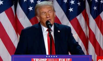 Donald Trump discursando em frente a bandeiras americanas, usando terno escuro e gravata vermelha, com o slogan 'Trump Will Fix It'.