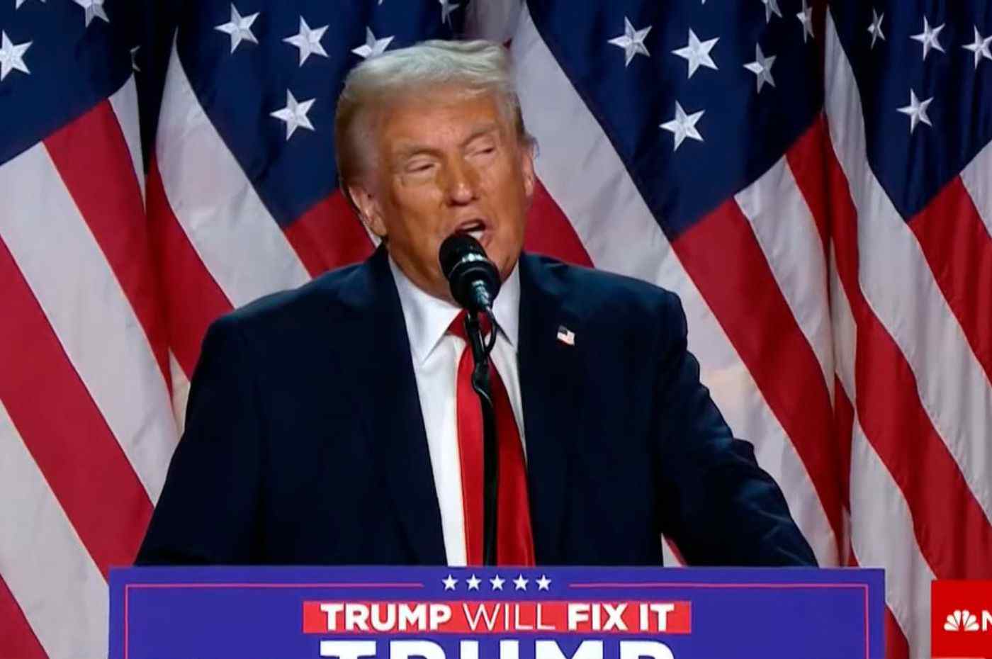 Donald Trump discursando em frente a bandeiras americanas, usando terno escuro e gravata vermelha, com o slogan 'Trump Will Fix It'.