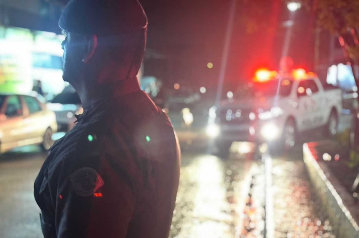 Agente de segurança em operação noturna em Jarinu, com viatura ao fundo e luzes vermelhas iluminando a rua para fiscalização.